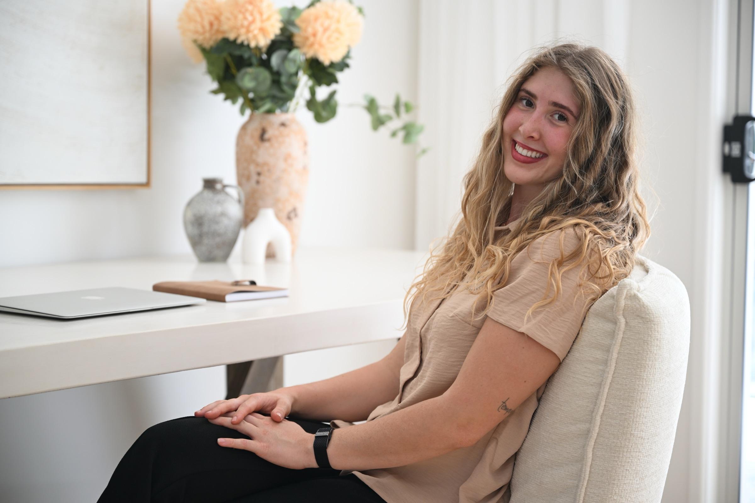 Bahli, founder of Bloom & Nurture, smiling at a white desk with a vase of flowers and a laptop