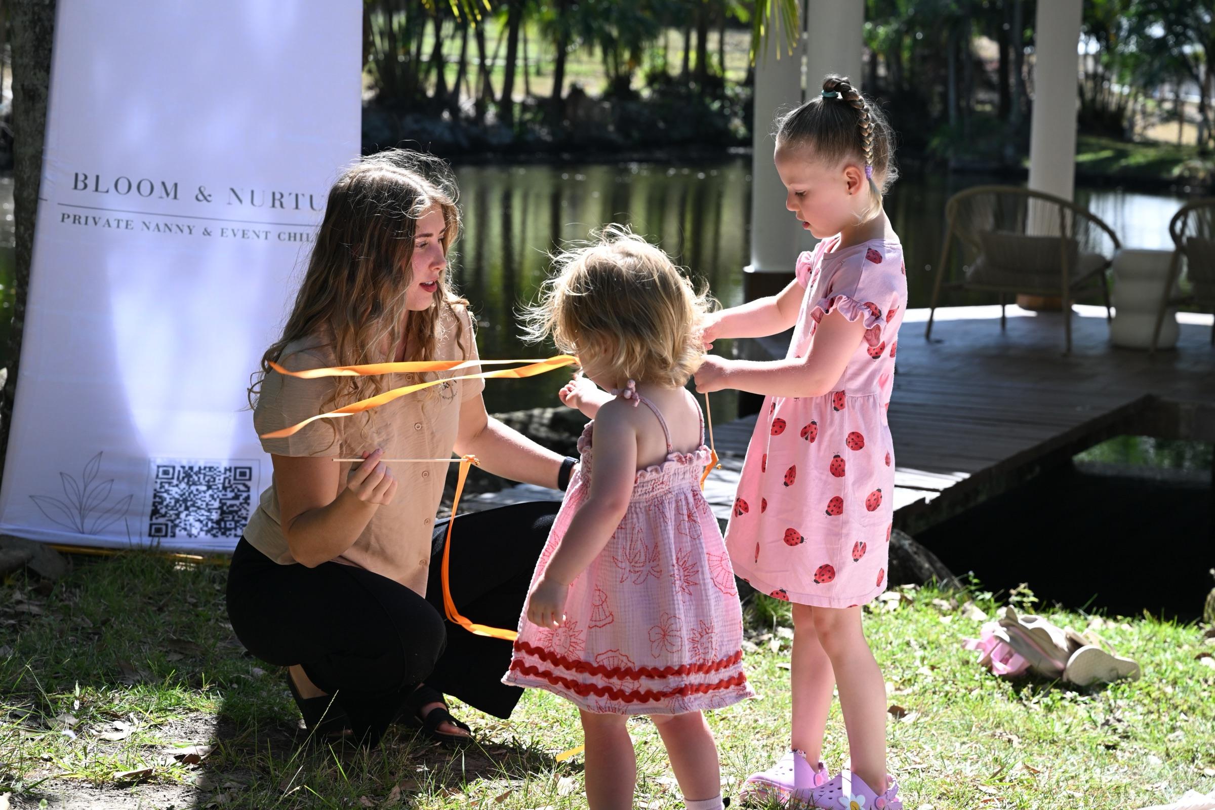 A nanny kneeling with a toddler and a young girl, holding an orange ribbon, in front of a Bloom & Nurture banner