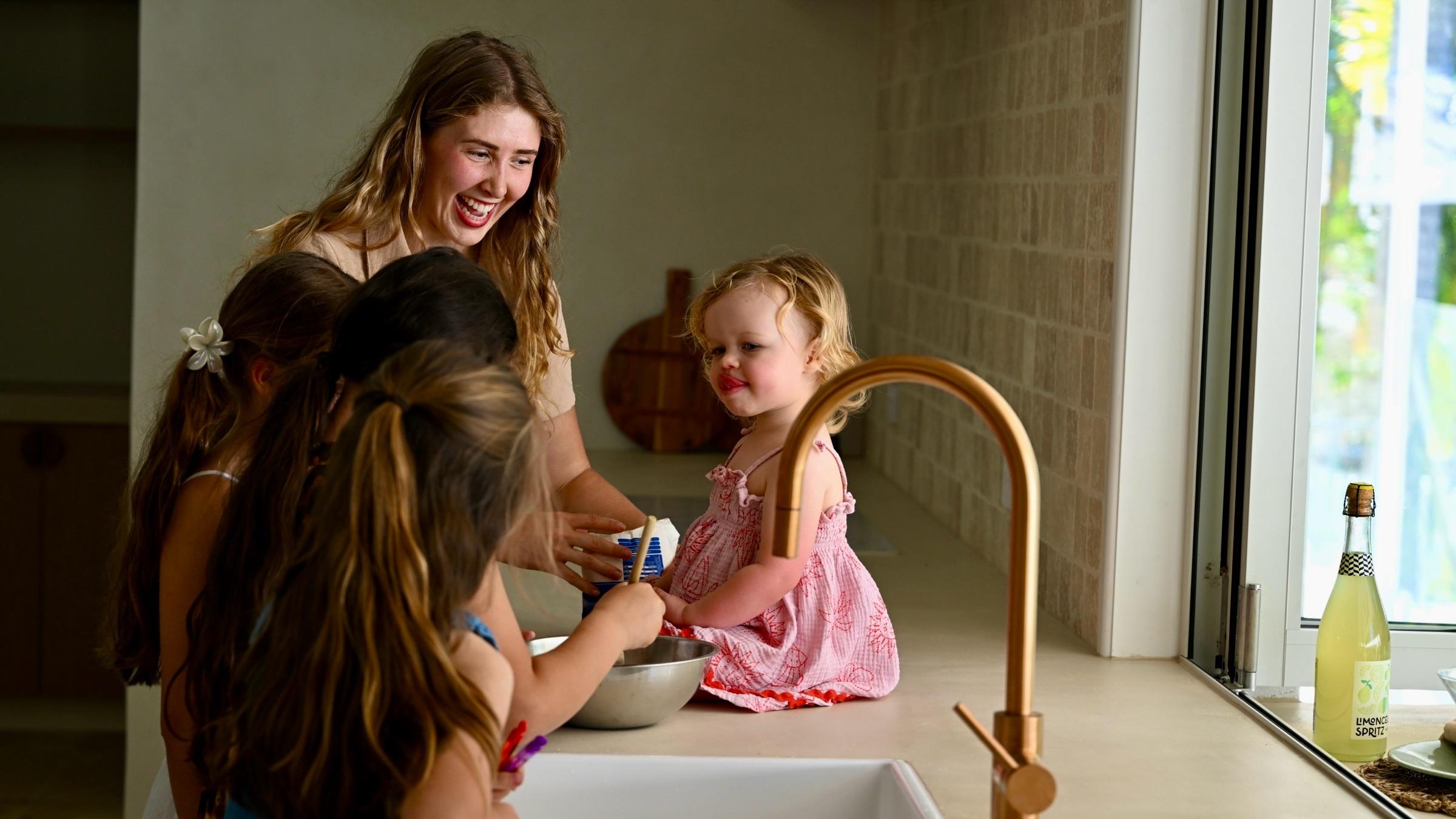 A nanny laughing in the kitchen as a toddler sits on the bench and two girls stir a mixing bowl