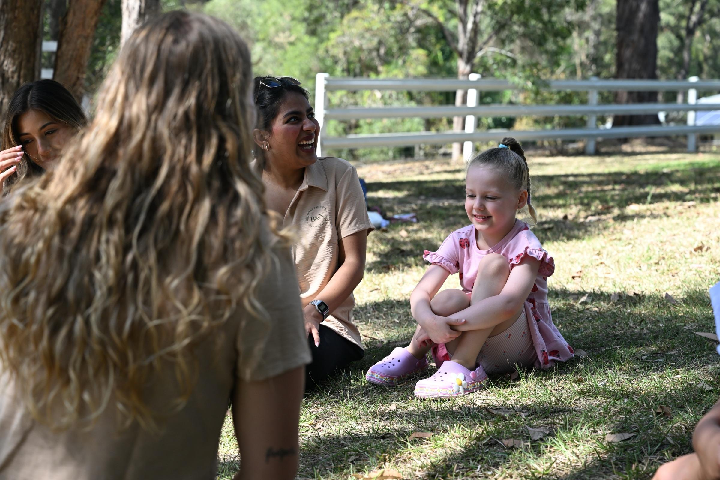A little girl smiling on the grass while carers around her laugh, at an outdoor event