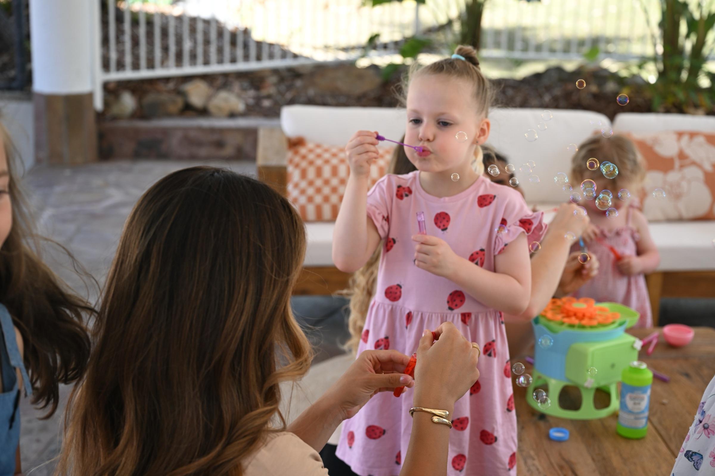 A little girl blowing bubbles at a wooden table beside a bubble machine, a carer’s hands helping in the foreground
