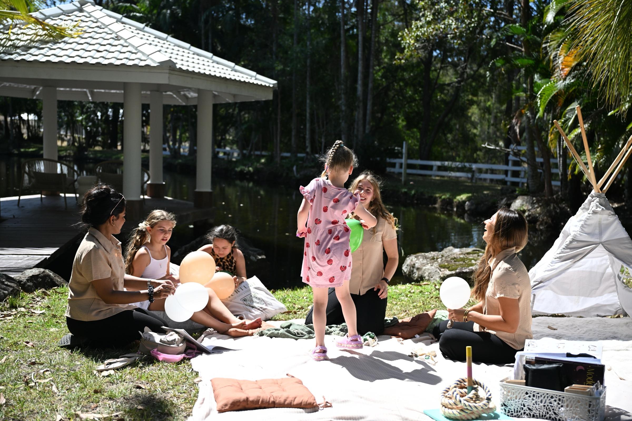 Carers and children playing with balloons on a picnic rug beside a lake