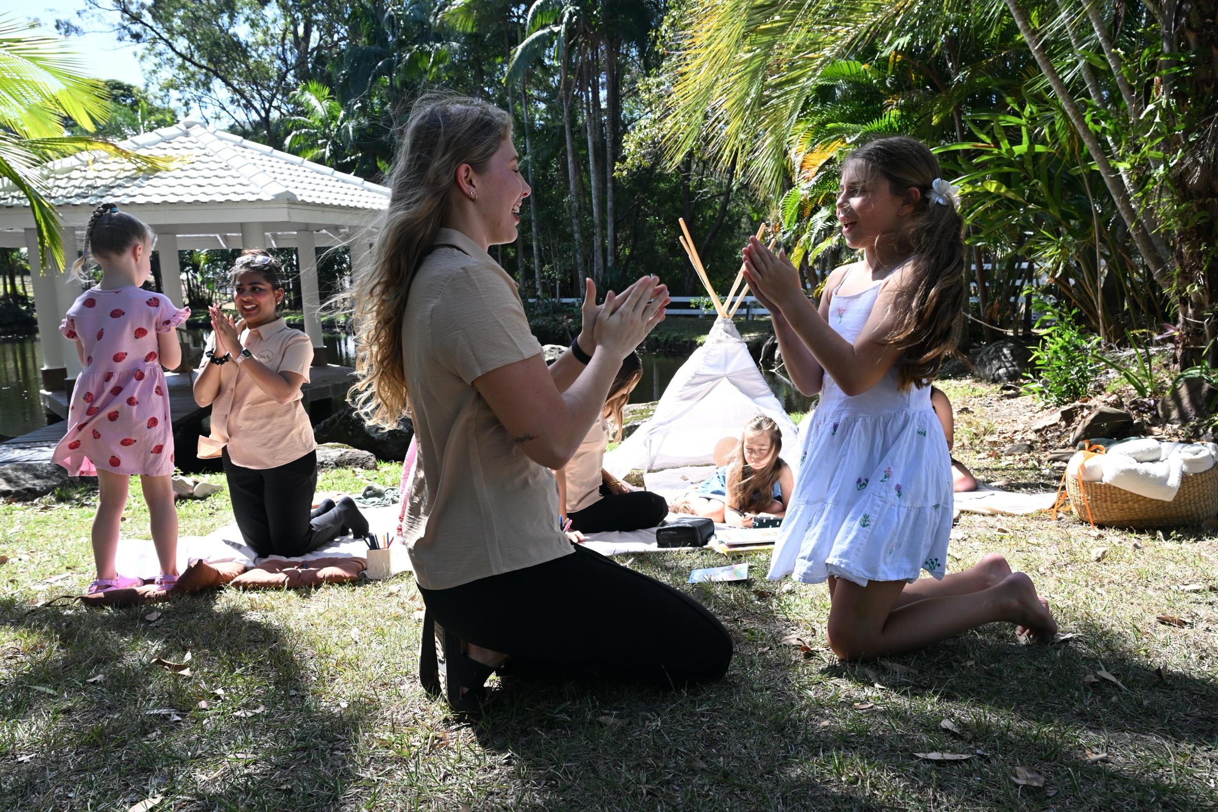 A carer kneeling on the grass clapping with a laughing girl, children and carers on a picnic rug behind them