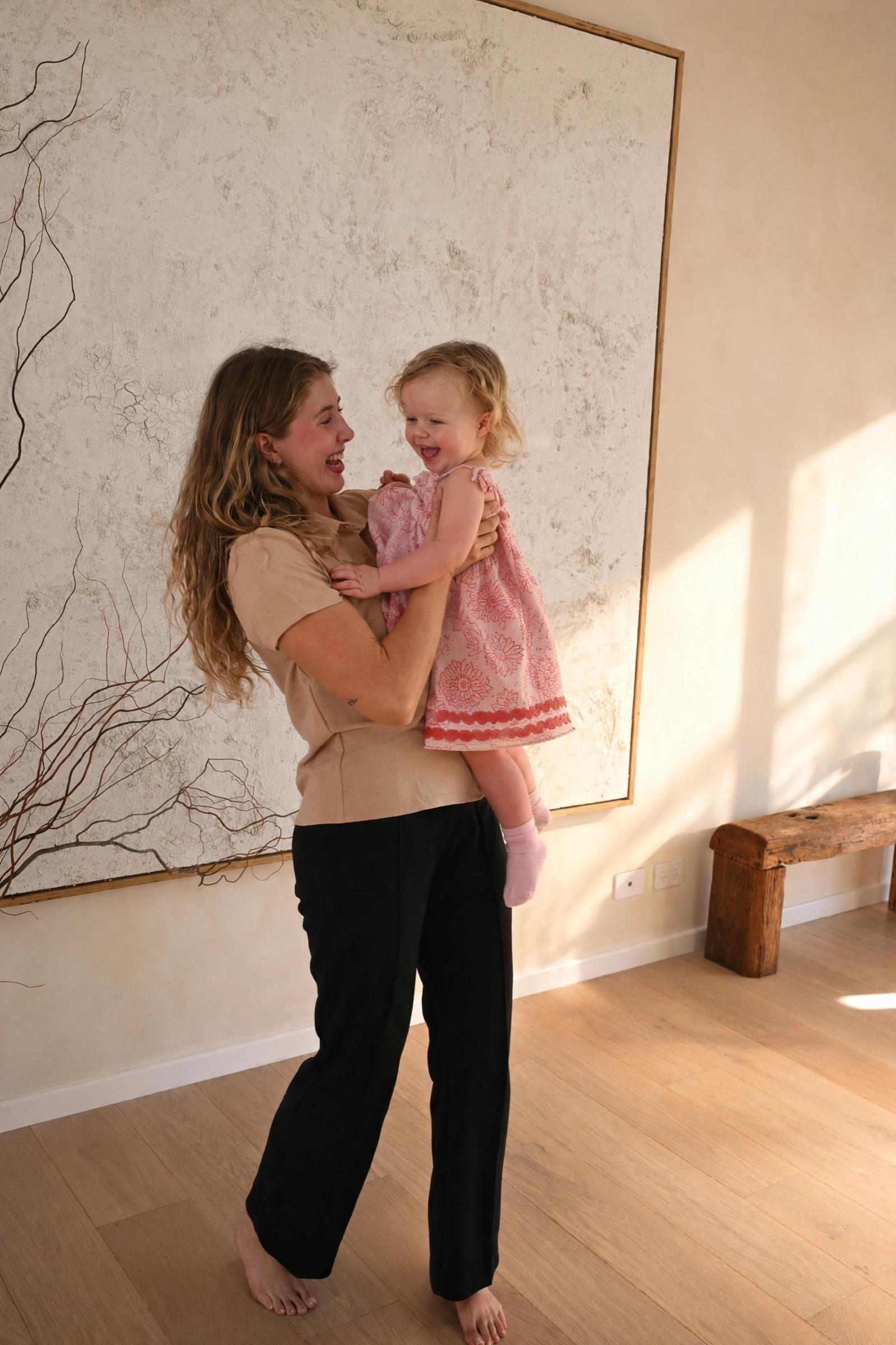 A nanny holding a laughing toddler in a bright, calm room with a large textured artwork behind them