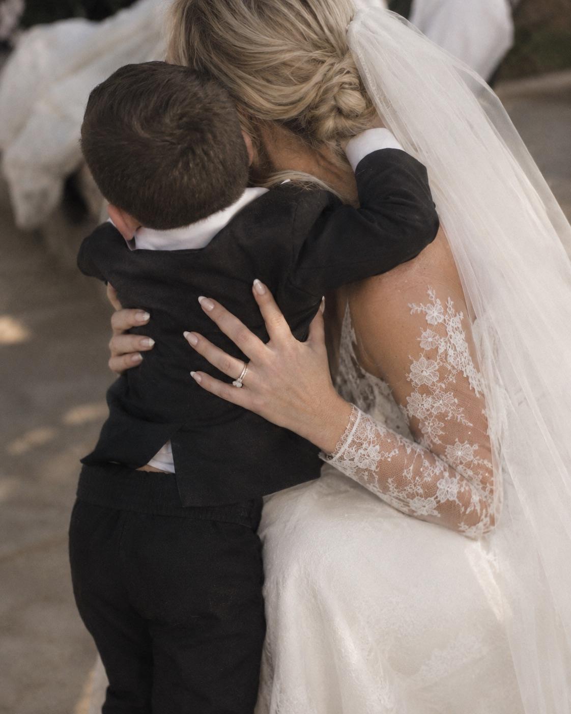 A little boy in a black waistcoat hugging a bride in a lace gown