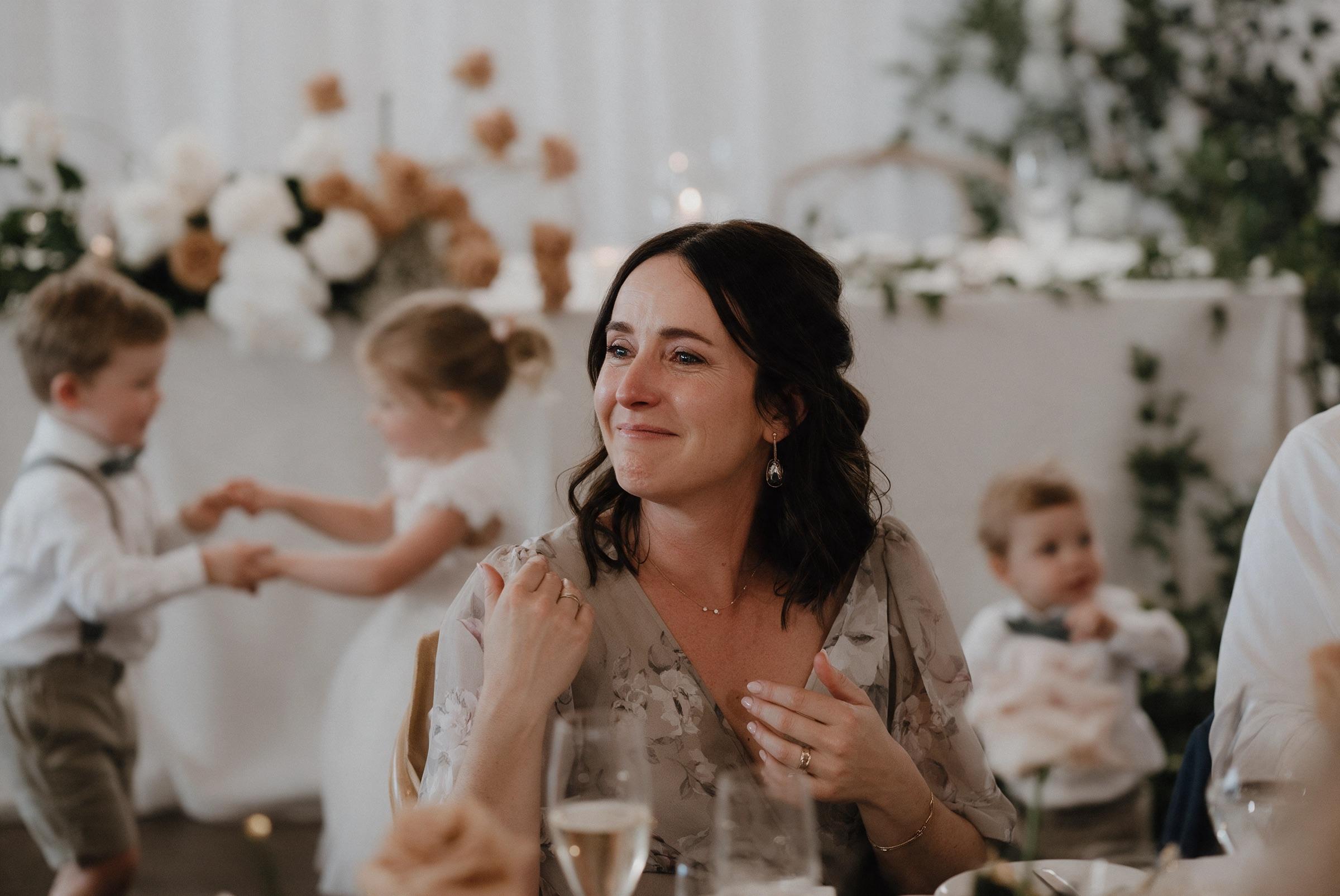 A guest smiling with tears in her eyes at a reception table while two children dance behind her