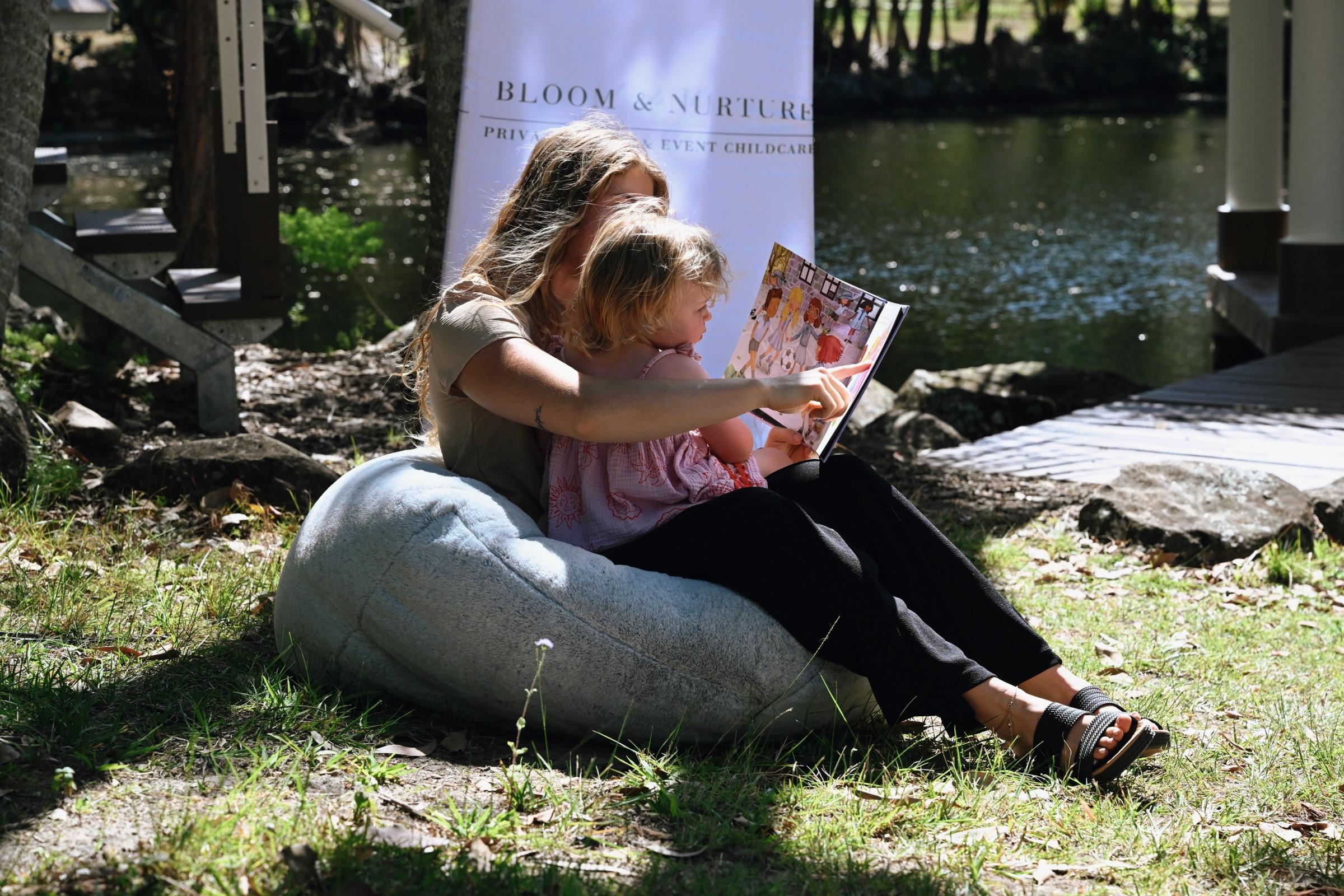 A carer reading a picture book to a toddler on a beanbag beside the water