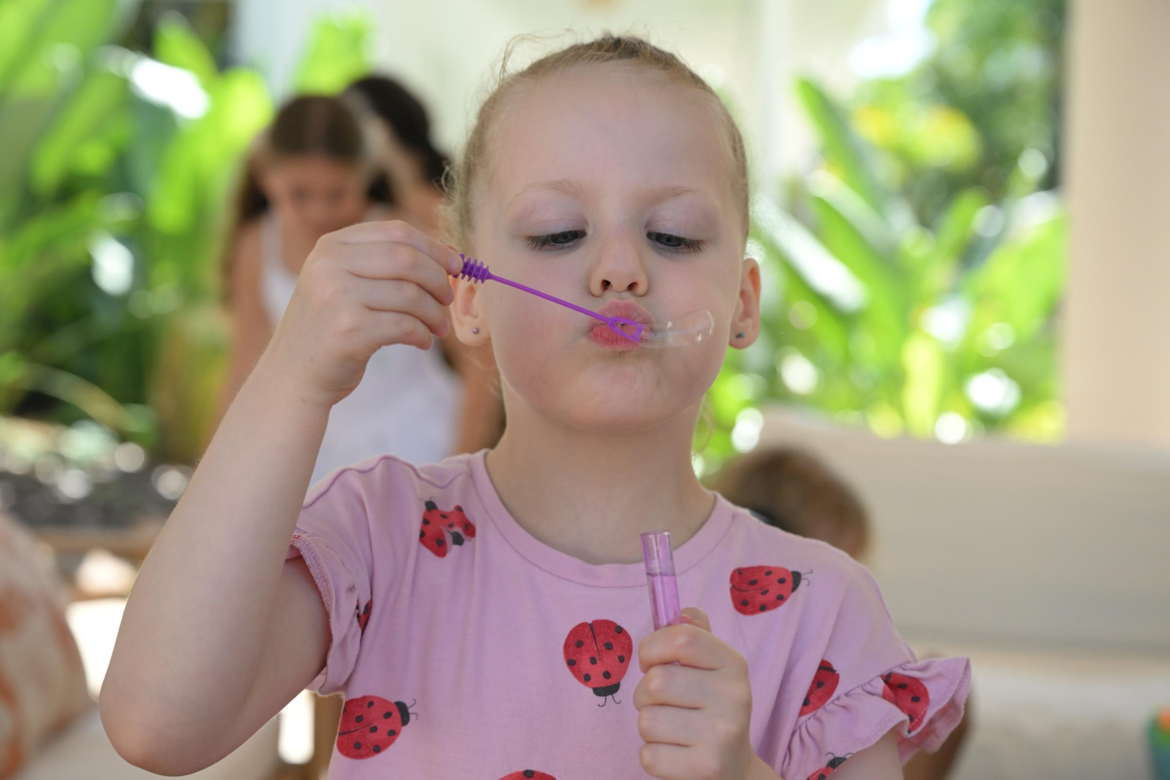 A little girl in a ladybird-print top blowing bubbles through a purple wand