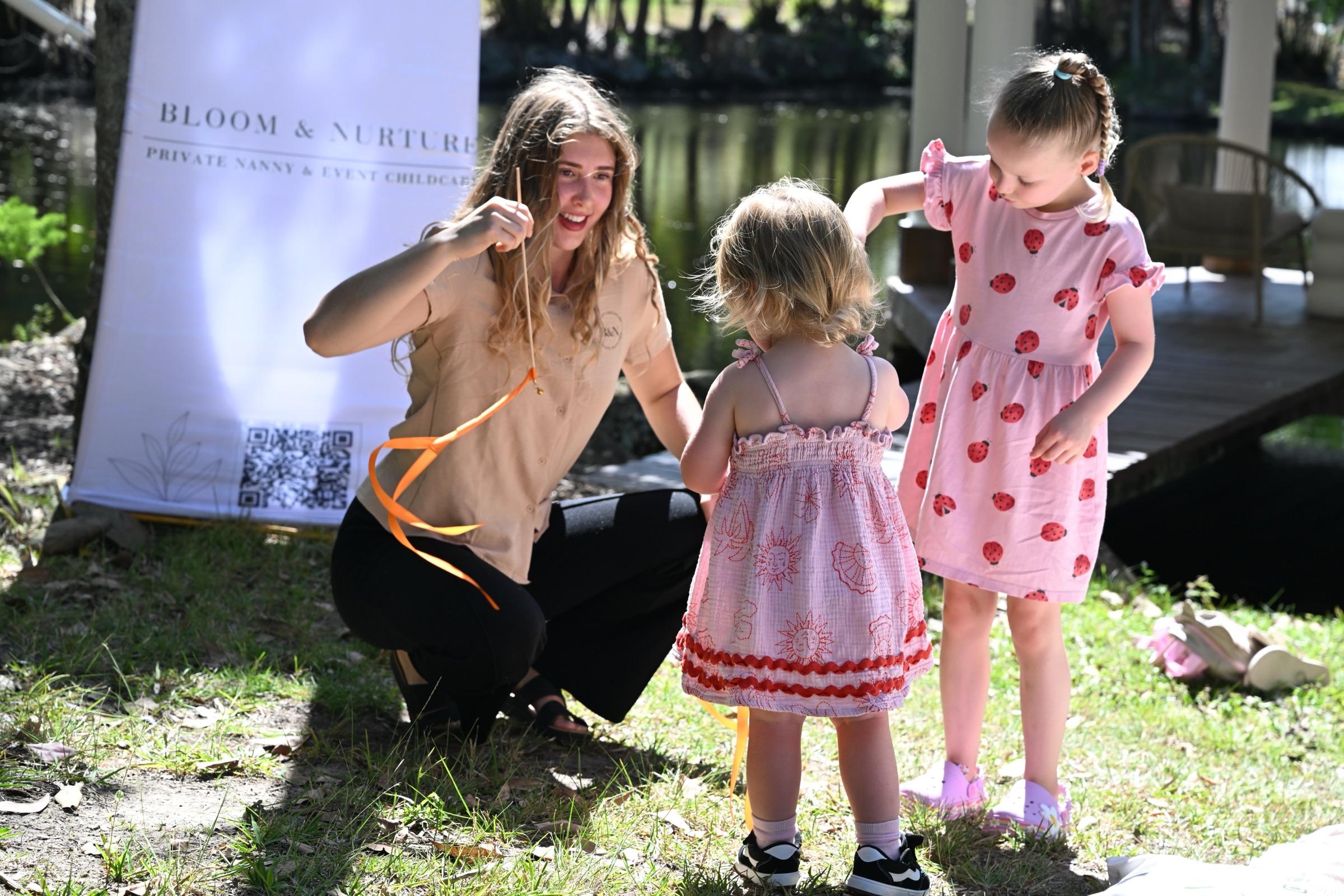 A nanny kneeling to hold an orange ribbon for a toddler and a young girl in front of a Bloom & Nurture banner