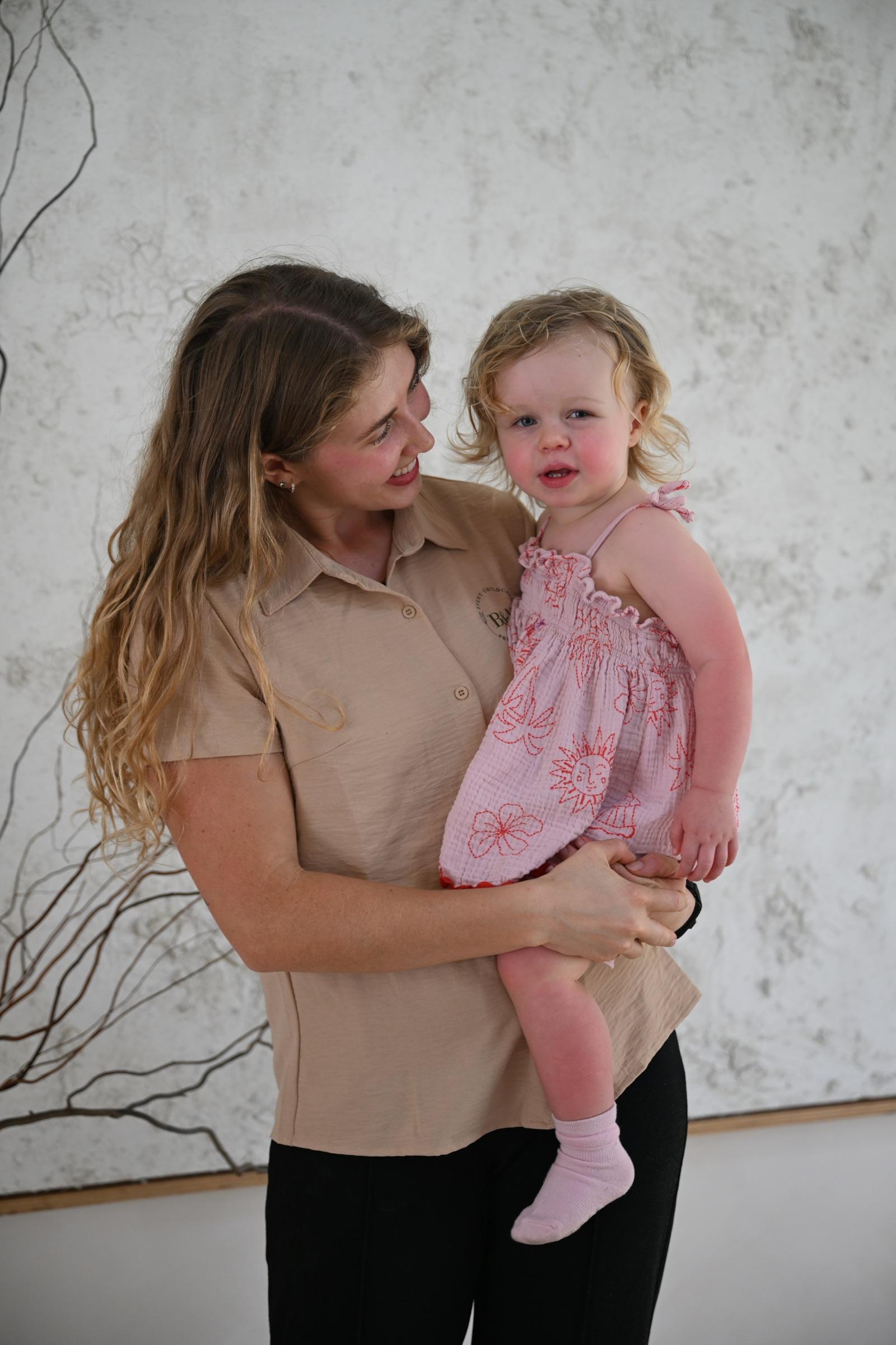 A nanny holding a toddler on her hip, smiling down at her, in front of a large textured artwork