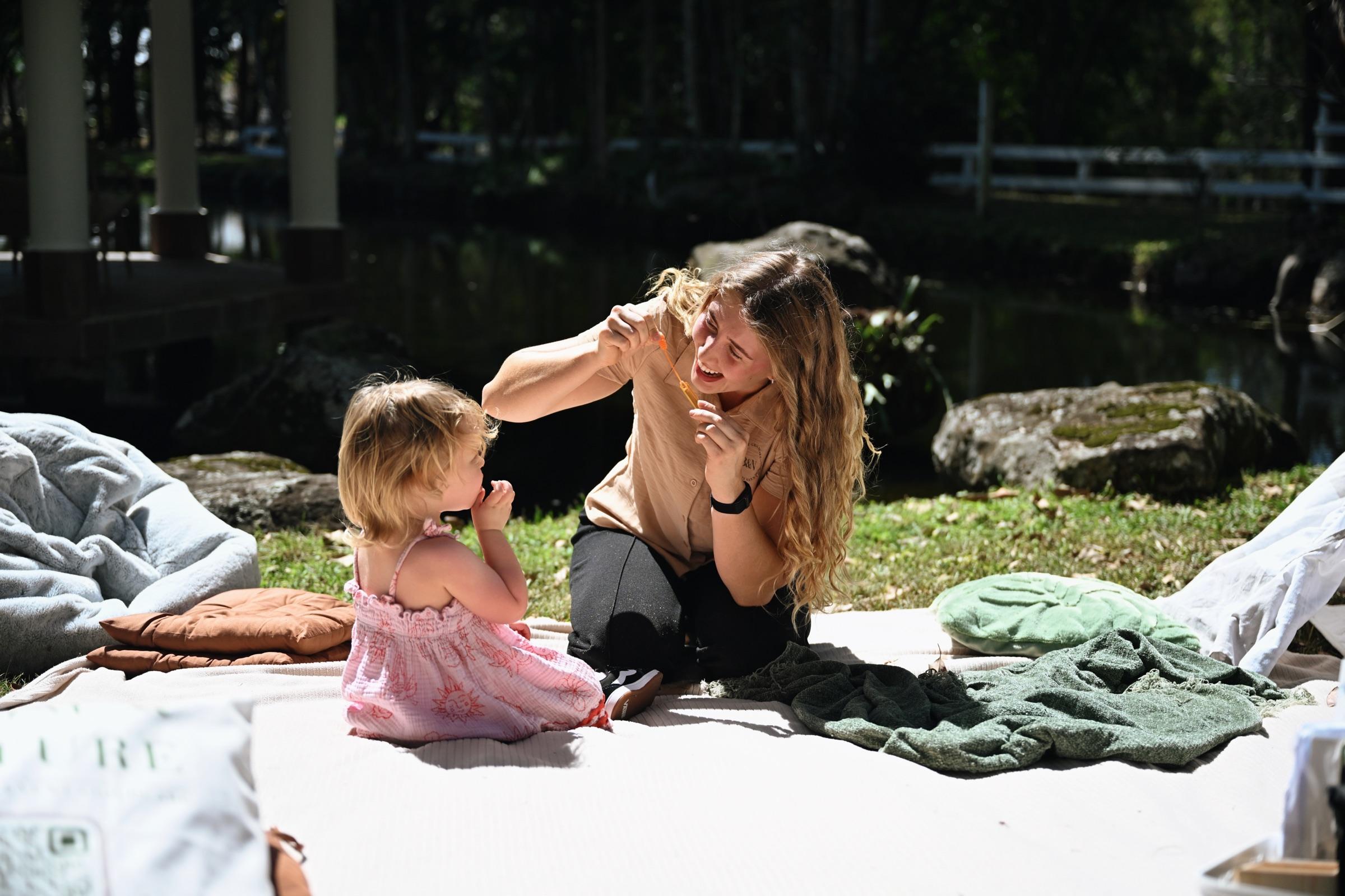 A carer playing with a toddler on a picnic rug beside a lake