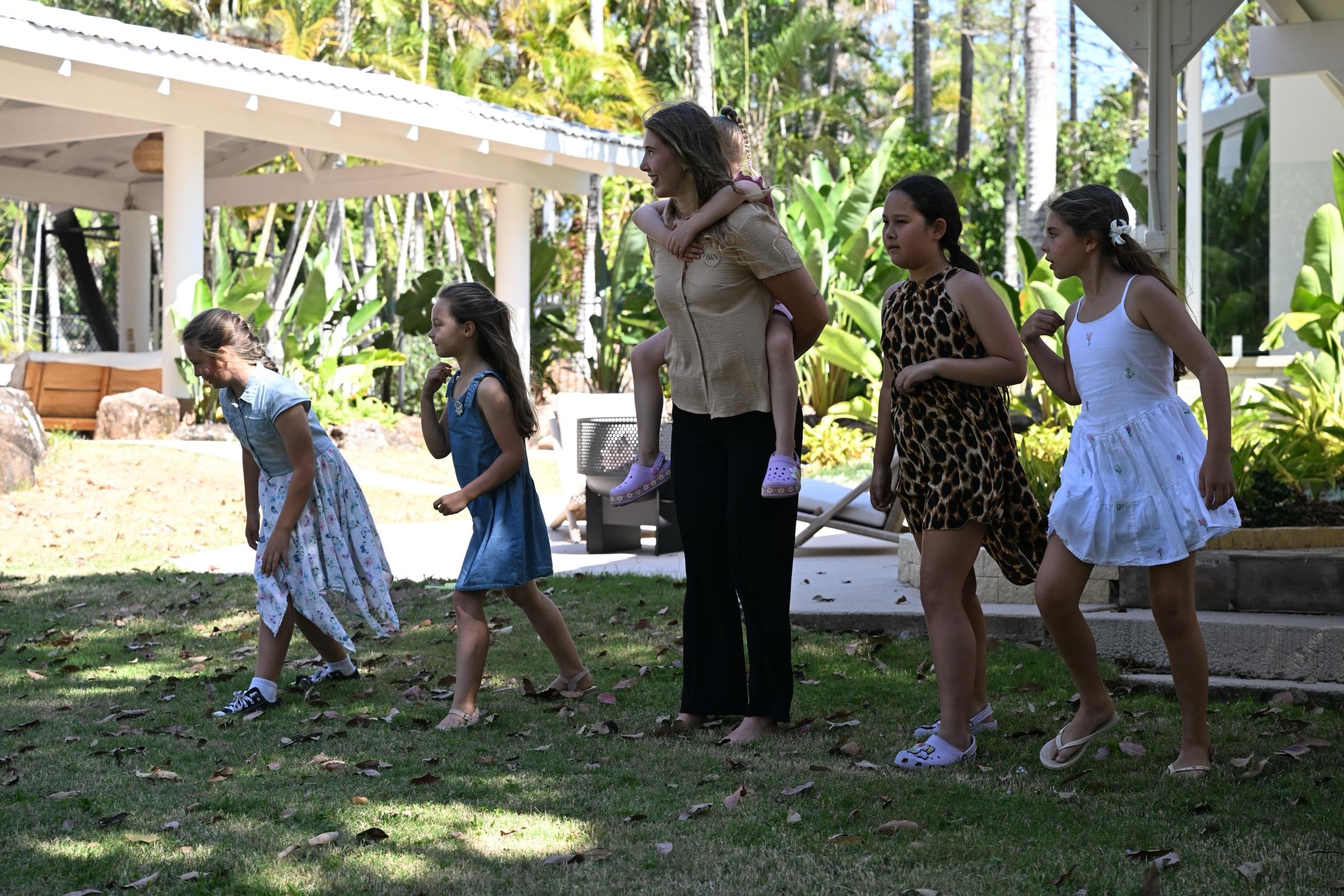 A carer carrying a toddler on her back while girls play a lawn game beside her