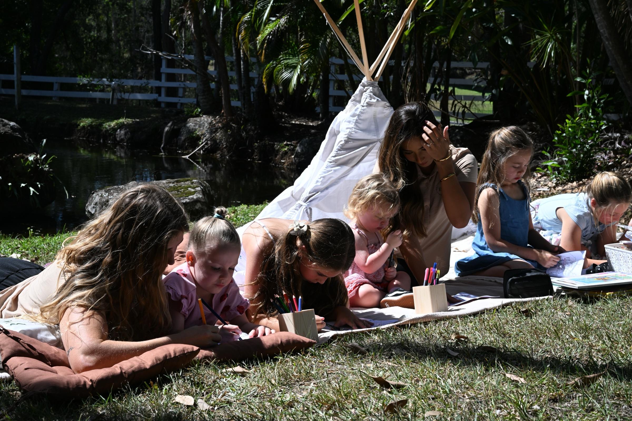 Children drawing with coloured pencils on a rug in the garden, carers beside them and a white tipi behind