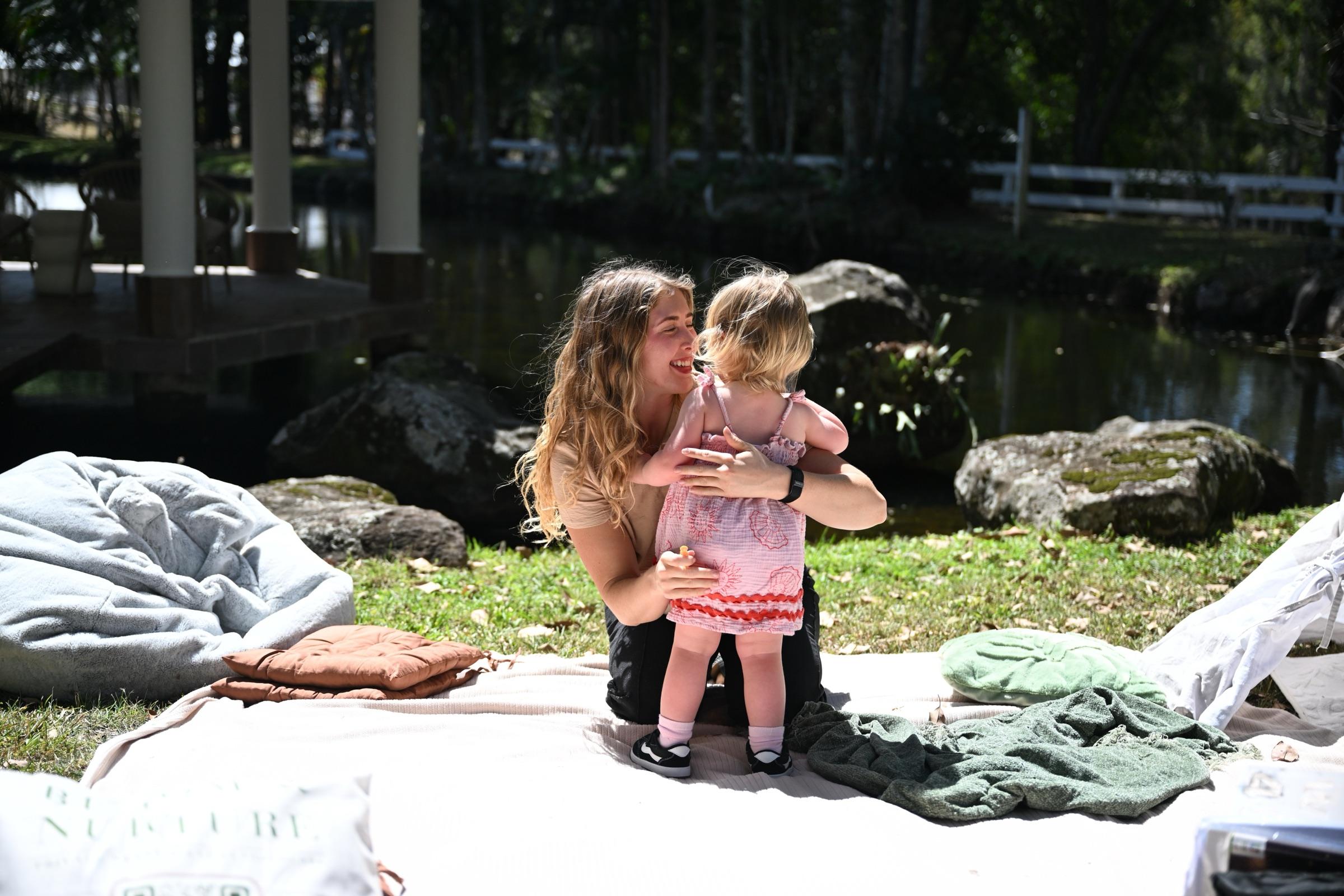 A carer crouched on a picnic rug, laughing as a toddler hugs her
