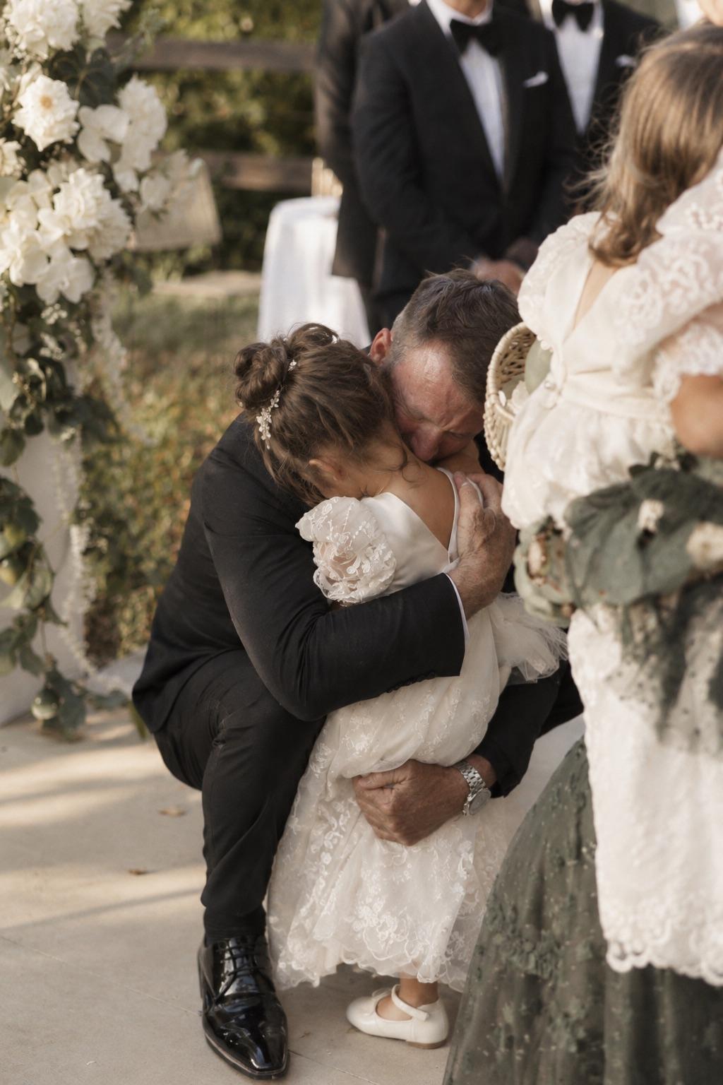 A little girl in a lace dress hugging a man in a suit at a wedding ceremony