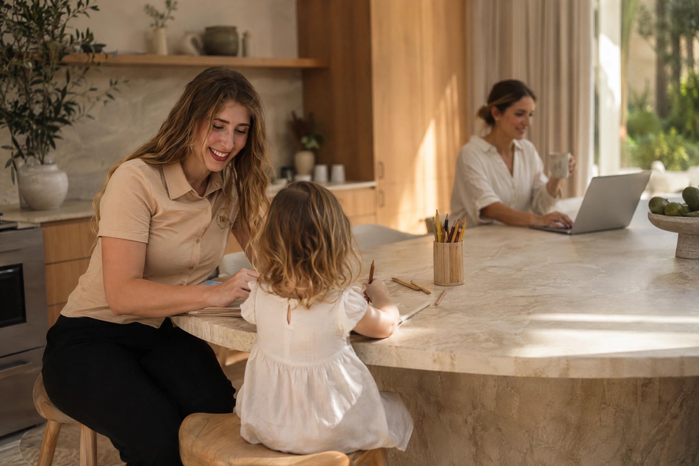 A nanny drawing with a toddler at a kitchen island while a parent works on a laptop behind them
